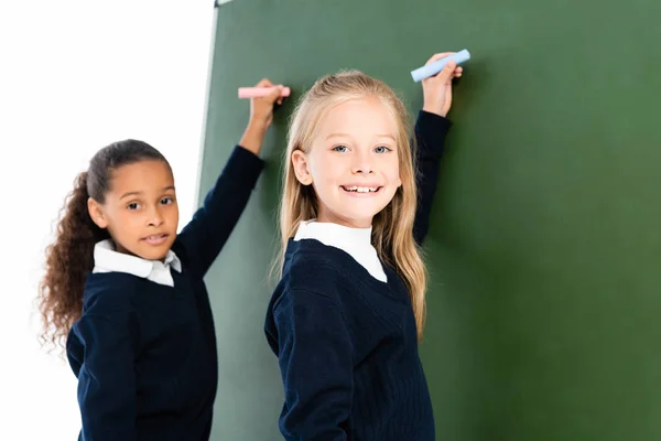 Zwei lächelnde multikulturelle Schulmädchen, die auf Kreidetafel schreiben, während sie in die Kamera schauen — Stockfoto