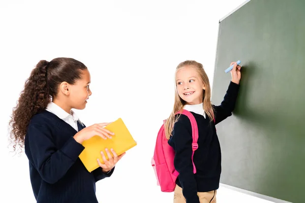 Zwei lächelnde Schulmädchen, die auf Klemmbrett schreiben und eine afrikanisch-amerikanische Klassenkameradin anschauen, die Buch isoliert auf weißem Papier hält — Stockfoto