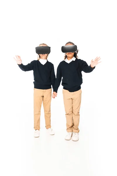 Ansicht von zwei multikulturellen Schulmädchen, die sich Händchen halten, während sie Virtual-Reality-Headsets auf weißem Hintergrund verwenden — Stockfoto