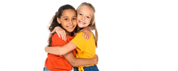 Foto panorámica de dos felices amigos multiculturales abrazándose mientras mira a la cámara aislada en blanco - foto de stock