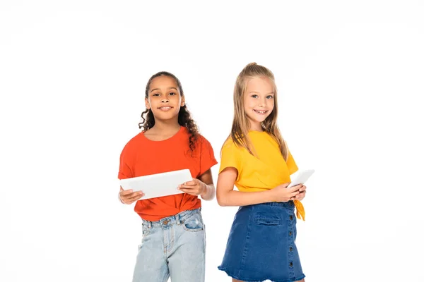 Dos amigos multiculturales sonrientes sosteniendo la tableta digital y el teléfono inteligente mientras sonríen a la cámara aislada en blanco - foto de stock