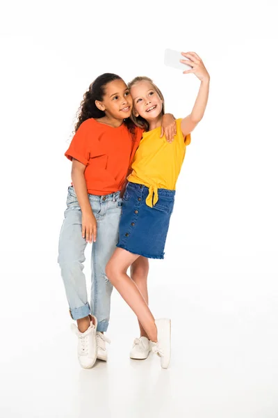 Full length view of happy african american child hugging smiling friend taking selfie with smartphone on white background — Stock Photo
