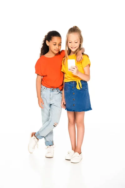 Vue pleine longueur de mignon afro-américain enfant étreignant ami en utilisant un smartphone sur fond blanc — Photo de stock