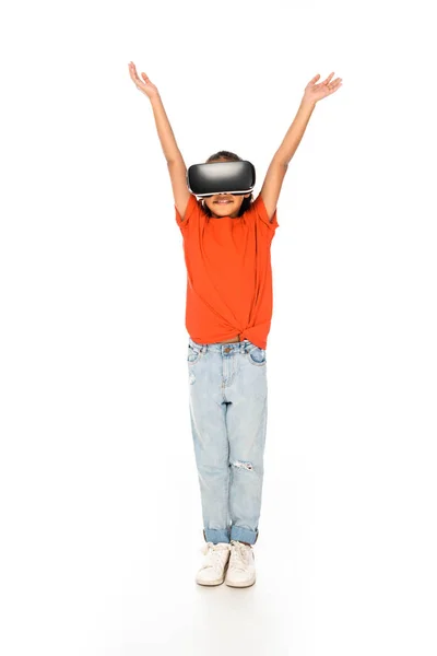 Vista completa de niño afroamericano sonriente de pie con las manos levantadas mientras usa auriculares de realidad virtual sobre fondo blanco - foto de stock