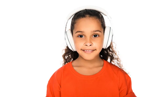 Adorable niño afroamericano sonriendo a la cámara mientras escucha música en auriculares aislados en blanco - foto de stock