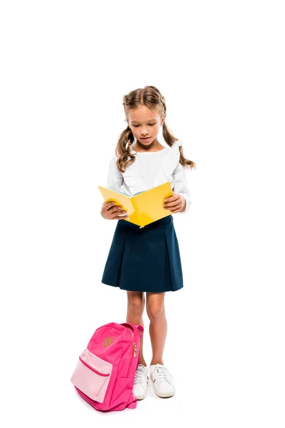 Bonito criança leitura livro e de pé perto rosa mochila isolada no branco — Fotografia de Stock