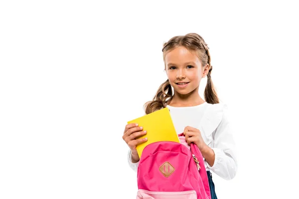 Fröhliches Kind legt Buch in rosa Rucksack, während es isoliert auf weiß steht — Stockfoto