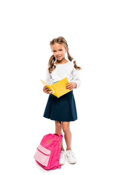 Libro de lectura infantil feliz y de pie cerca de la mochila rosa aislado en blanco - foto de stock