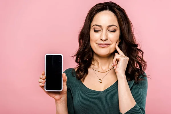 Щаслива жінка торкається обличчя і тримає смартфон з порожнім екраном ізольовано на рожевому — Stock Photo