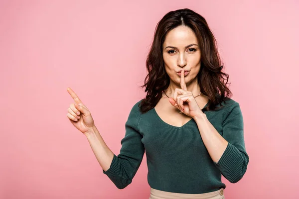 Attractive woman showing hush sign and pointing with finger isolated on pink — Stock Photo