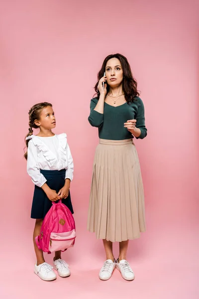 Filha bonito segurando mochila e olhando para a mãe falando no smartphone em rosa — Fotografia de Stock