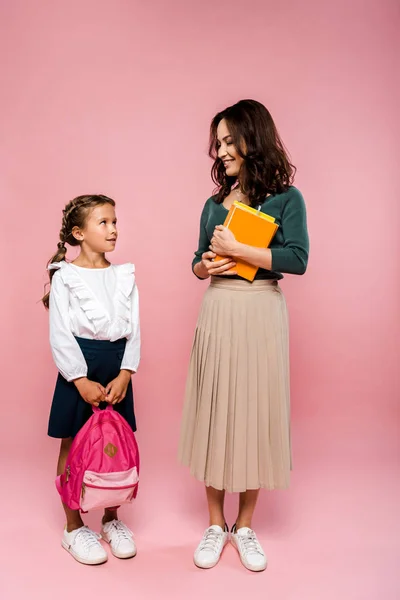 Feliz madre sosteniendo libros y de pie cerca de linda hija con mochila en rosa - foto de stock