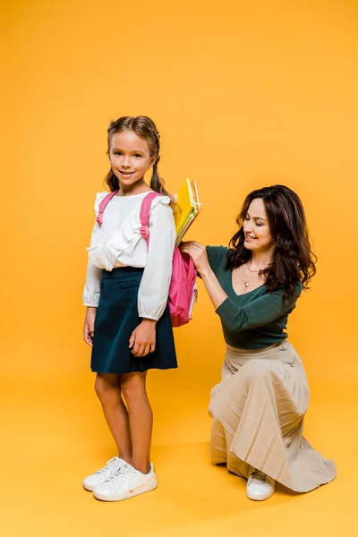 Attractive mother putting books in backpack of schoolgirl on orange — Stock Photo