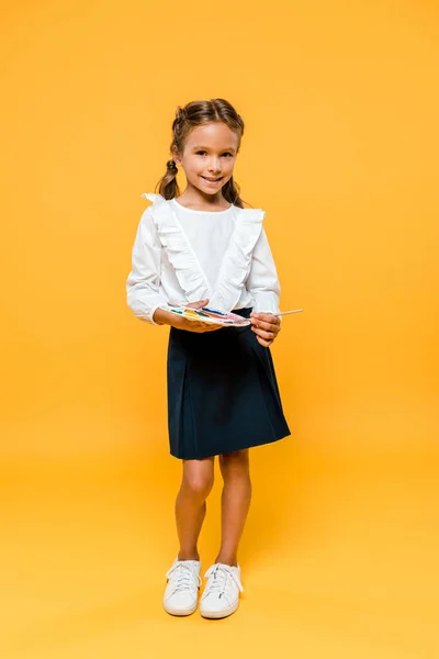 Cheerful schoolgirl holding palette and paintbrush on orange — Stock Photo