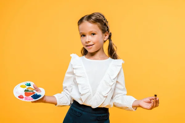 Fröhliches Schulmädchen mit Palette und Pinsel isoliert auf orange — Stockfoto