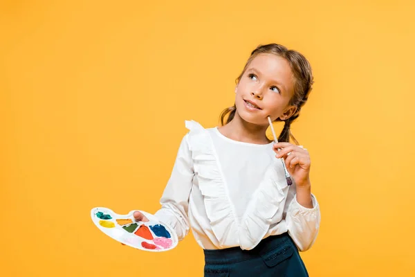 Pensive schoolkid holding palette and paintbrush isolated on orange — Stock Photo