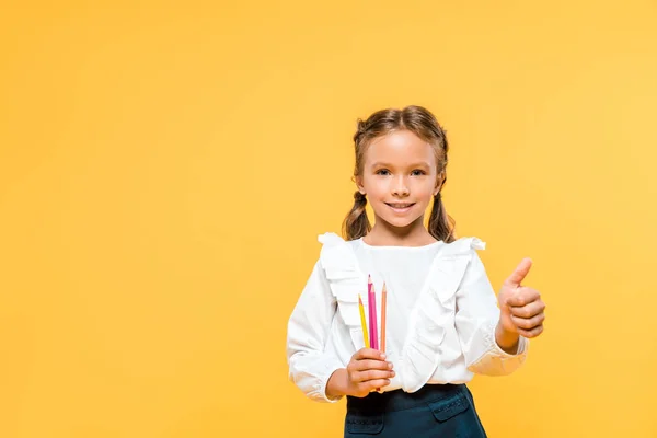 Heureux écolier tenant crayons de couleur et montrant pouce vers le haut isolé sur orange — Photo de stock