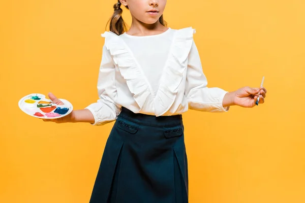 Cropped view of schoolkid  holding palette and paintbrush isolated on orange — Stock Photo