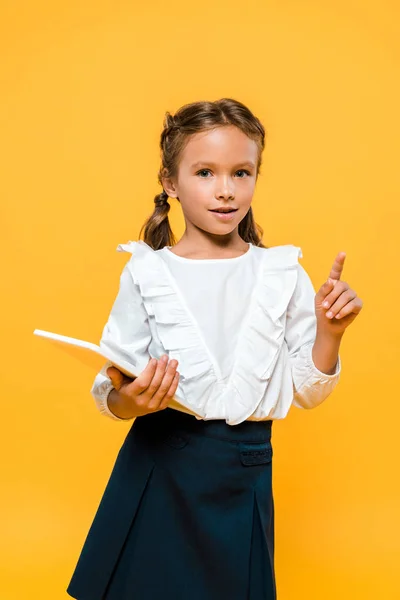 Happy schoolkid holding book and pointing with finger isolated on orange — Stock Photo