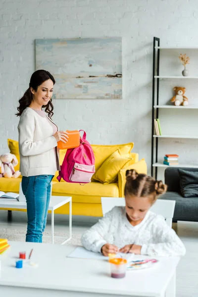 Selective focus of happy mother putting lunch box in backpack of daughter — Stock Photo
