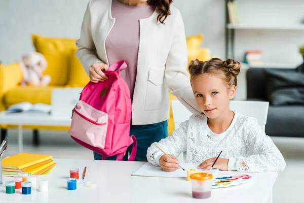 Corte vista de mãe segurando mochila perto de bonito filha pintura em casa — Fotografia de Stock