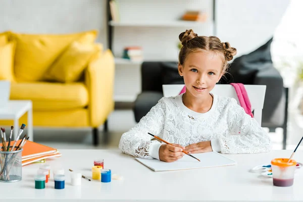 Heureux enfant tenant pinceau près des pots de gouache et du papier sur la table — Photo de stock