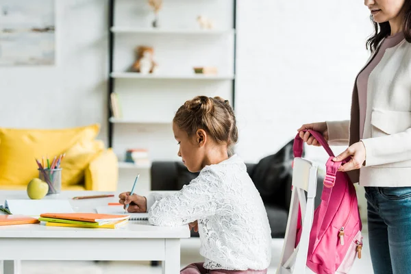 Cropped view of mother holding pink backpack near daughter drawing at home — Stock Photo