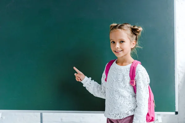 Cheerful kid smiling while pointing with finger at green chalkboard — Stock Photo