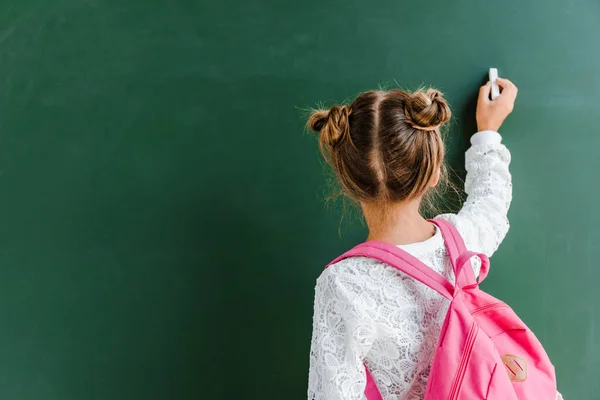 Vista trasera de un niño en edad escolar sosteniendo tiza cerca de pizarra en verde - foto de stock