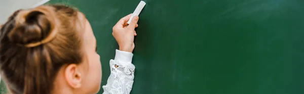 Panoramic shot of schoolchild holding chalk near chalkboard on green — Stock Photo