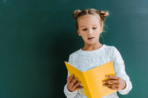 Surpris écolière lecture livre jaune sur vert — Photo de stock