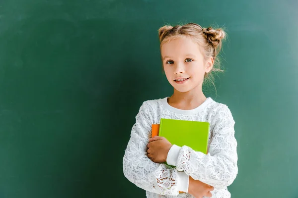 Lächelndes Schulmädchen, das lächelt, während es mit Büchern auf Grün steht — Stockfoto