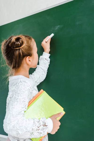 Niedliche Schulkind hält Kreide und Bücher in der Nähe der grünen Tafel — Stockfoto