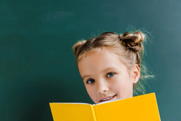 Criança feliz sorrindo enquanto segurando livro amarelo no verde — Fotografia de Stock