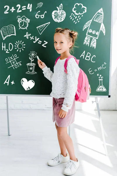 Happy kid standing with backpack and holding chalk near drawing on green chalkboard — Stock Photo