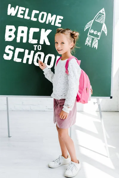 Cute schoolgirl standing with backpack and holding chalk near welcome back to school lettering on green chalkboard — Stock Photo