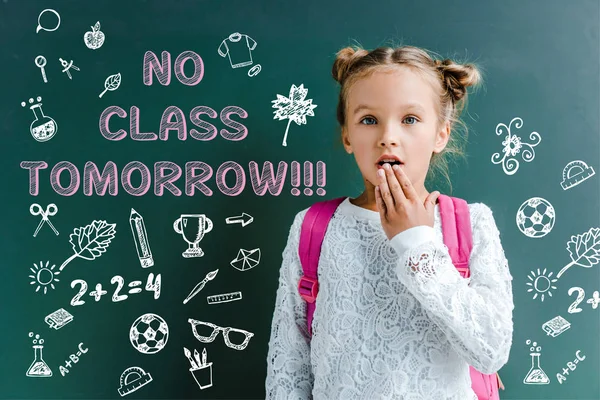 Surprised schoolgirl covering mouth near chalkboard with no class tomorrow lettering on green — Stock Photo