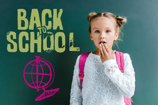 Scolaretta sorpresa che copre la bocca vicino alla lavagna con le lettere di ritorno a scuola sul verde — Foto stock