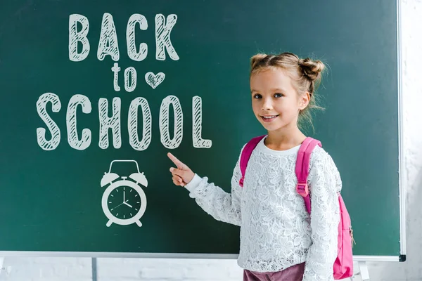 Criança alegre sorrindo ao apontar com o dedo para trás para a escola lettering no quadro verde — Fotografia de Stock