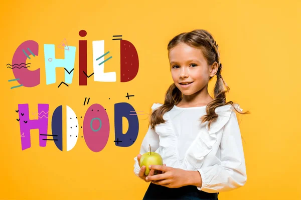 Niño de escuela feliz sosteniendo sabrosa manzana cerca de letras de la infancia en naranja - foto de stock