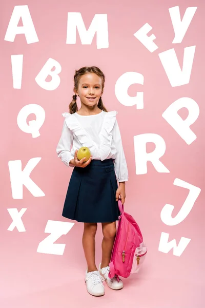 Happy kid holding tasty and green apple and backpack near letters on pink — Stock Photo