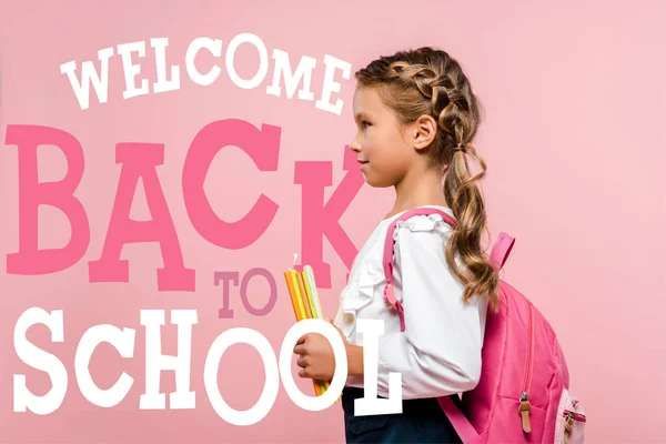 Side view of happy kid holding books while standing with backpack near welcome back to school lettering on pink — Stock Photo