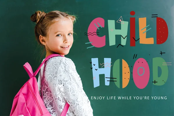 Glückliches Schulkind lächelt, während es mit Rucksack in der Nähe des Schriftzugs auf Grün steht — Stockfoto