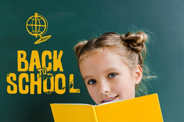 Enfant heureux souriant tout en tenant livre jaune près de retour à l'école lettrage sur vert — Photo de stock