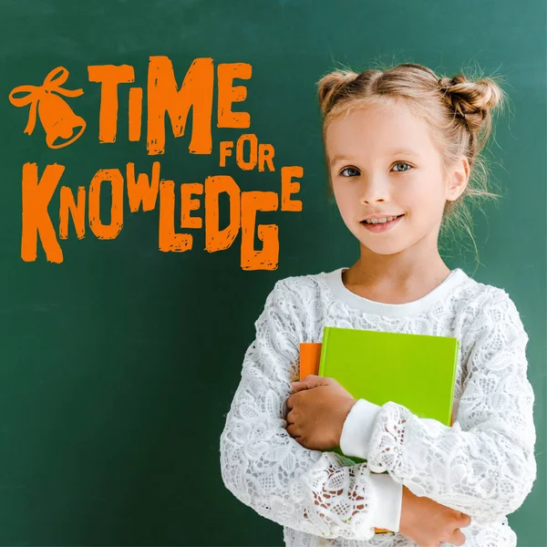 Happy schoolgirl smiling while standing with books near time for knowledge lettering on green — Stock Photo