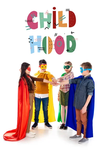 Happy kids in superhero costumes and masks stacking hands near childhood lettering  on white — Stock Photo