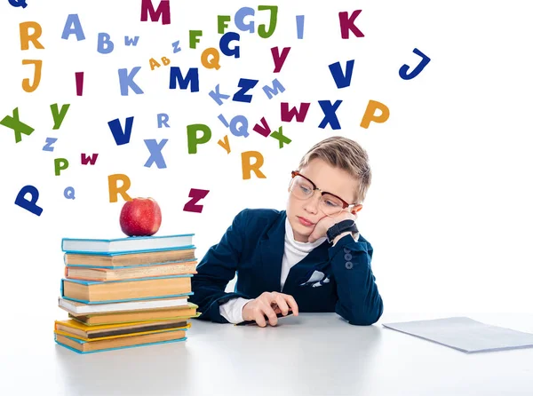Menino de escola triste em óculos sentado na mesa com livros e maçã perto de letras coloridas em branco — Fotografia de Stock