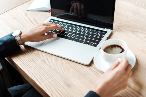 Cropped view of businesswoman typing on laptop with blank screen near cup of coffee on table — Stock Photo