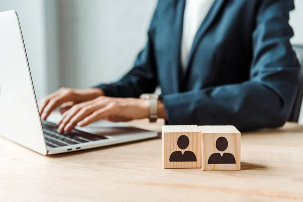 Cropped view of businesswoman typing on laptop near wooden cubes with human shapes — Stock Photo