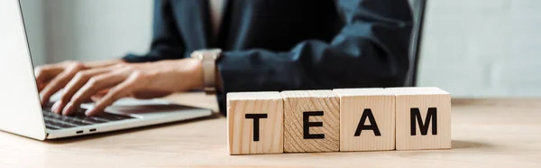 Panoramic shot of businesswoman typing on laptop near wooden cubes with team letters — Stock Photo